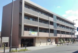介護施設の建物外観。茶色のタイル張りの5階建て建物で、1階に玄関・駐車場があり、バルコニー付きの居住階が上部に広がっている。